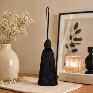May include: A black decorative tassel featuring a smooth, rounded top cap and a long, dense fringe of fine, silky threads. The tassel is suspended by a matching black braided cord loop, creating a clean, vertical silhouette for home decor.