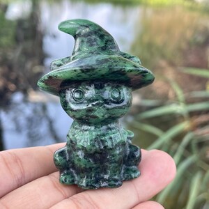 May include: A small, carved stone figurine of a cat wearing a pointed witch hat. The sculpture is crafted from a dark green mineral with black mottled patterns throughout, featuring large, round eyes and a detailed collar around the neck area.
