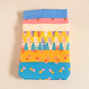 May include: A stack of seven folded fabric pieces featuring various patterns including blue with speckles, solid pink with small orange fruit, cream with colorful speckles, ice cream cones, yellow with cherries, and blue with small cherries.