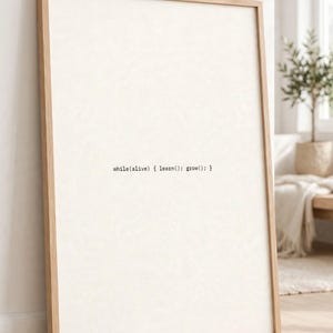 May include: A rectangular framed wall art piece featuring a light cream background with the black typewriter-style text while(alive) { learn(); grow(); } centered in the middle. The frame is made of light-colored wood with a simple, clean profile.