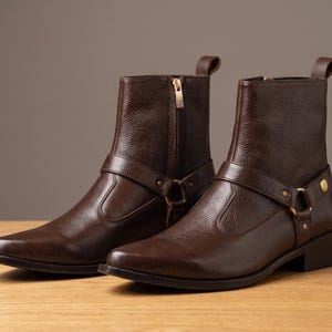 May include: A pair of dark brown ankle boots featuring a textured leather finish, a side zipper closure, and a decorative harness strap with a gold-toned metal ring and stud detail. The boots have a pull tab at the heel and a low block heel.