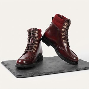 Pode incluir: Um par de botins em bordeaux escuro com acabamento brilhante, atacadores castanhos e ilhós de metal dourado. As botas têm um colarinho almofadado em tecido vermelho profundo a condizer, biqueira redonda e sola grossa de borracha preta.
