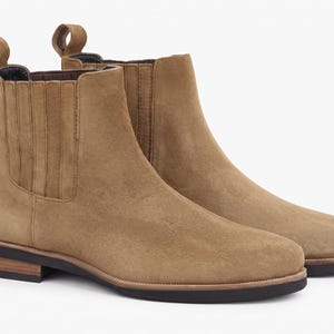May include: A pair of tan suede Chelsea boots featuring elastic side panels, a pull tab at the heel, and a low-profile dark brown sole with a stacked wood-look heel detail. The boots have a rounded toe and a smooth, uniform finish throughout.