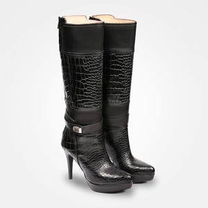 May include: A pair of black knee-high boots featuring a textured crocodile-embossed pattern on the shaft and foot. The boots include a smooth leather strap across the ankle with a metallic buckle, a high stiletto heel, and a platform sole.