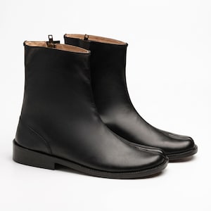 May include: A pair of black leather ankle boots featuring a distinct split-toe design, a low block heel, and a vertical zipper closure located at the inner side of the shaft. The boots have a smooth finish and a beige interior lining.