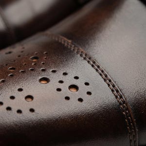 May include: A dark brown leather shoe featuring a cap toe design with decorative brogue perforations arranged in circular patterns. The leather has a textured finish with visible double-row stitching along the seam of the toe cap.