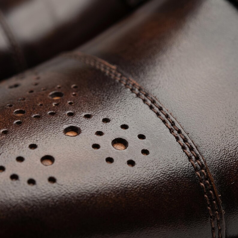 May include: A dark brown leather shoe featuring a cap toe design with decorative brogue perforations arranged in circular patterns. The leather has a textured finish with visible double-row stitching along the seam of the toe cap.
