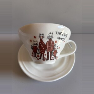 May include: A white ceramic teacup and saucer set featuring a brown illustration of four cats wearing sweaters. The text THE CAT'S FAMILY is printed in black on the side of the cup. The cup has a curved handle and the saucer has a ridged edge design.