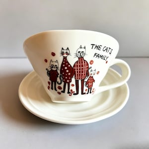 White porcelain cup and matching saucer with red and black cat-family illustration.