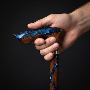 Puede incluir: Un bastón que presenta un mango y un eje elaborados con una combinación de madera marrón oscura y resina azul arremolinada. El mango tiene una forma curva y ergonómica, y una banda metálica separa el mango del eje vertical de madera.