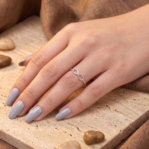 May include: A thin silver-toned ring featuring an infinity symbol design centred on the band. The ring is worn on the ring finger, showcasing a delicate metallic construction with a polished finish that forms a continuous loop shape.