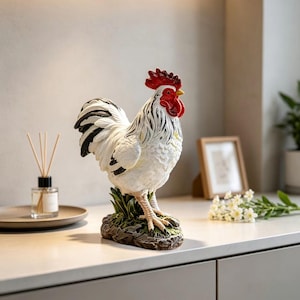 May include: A decorative rooster figurine featuring white plumage with black accents on the wings and tail, a bright red comb and wattle, and a yellow beak. The rooster stands on a textured base resembling a patch of green grass and rocky ground.