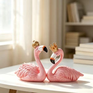 Puede incluir: Dos figuras de flamencos rosas frente a frente, cada una con una pequeña corona dorada. Los flamencos tienen plumas texturizadas, ojos cerrados y picos con puntas negras. Las figuras están colocadas una al lado de la otra.