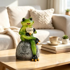 Op de afbeelding: Een groen kikkerbeeldje zittend op een grijze rots, met een kleine witte mok met het opschrift "coffee". Op de rots staat de tekst "Coffee gets me JUMPIN". De kikker heeft zwarte accenten op de voeten en handen met goudkleurige uiteinden.