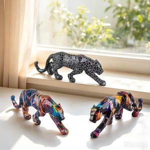 Peut inclure: Trois figurines de panthères en position de rôdeur. L'une est noire avec un motif craquelé blanc. Une autre présente un motif abstrait multicolore avec des stries bleues, rouges et jaunes. La troisième a un design géométrique vibrant.