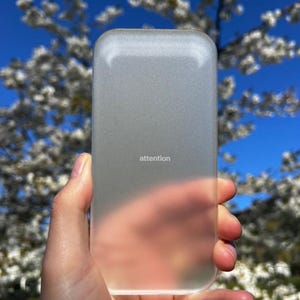 Peut inclure: Une coque de téléphone rectangulaire, semi-transparente et givrée avec des coins arrondis. Le mot "attention" est imprimé en petites lettres minuscules blanches sans empattement au centre de la coque, qui a une texture mate lisse.