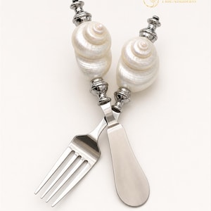 May include: A silver-toned fork and spreader set featuring handles crafted from white, spiraled seashells. The metal components are polished, and the top of the handles are accented with silver-toned finials. The text TideAndBrass is visible.