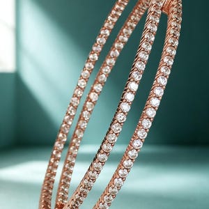 May include: A rose gold hinged bangle bracelet featuring two parallel rows of round, clear, faceted stones set in a twisted metal design. The bracelet has a secure clasp mechanism at the top and a polished metallic finish throughout the structure.