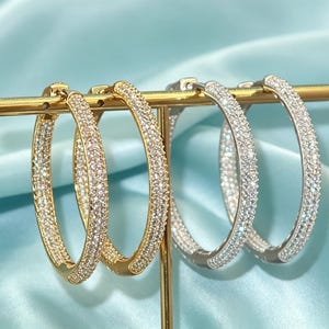 May include: Two pairs of hoop earrings are displayed on a gold-toned T-bar stand. One pair is gold-toned and the other is silver-toned. Both pairs feature a pavé-set design with numerous small, clear, sparkling stones covering the entire outer surface.