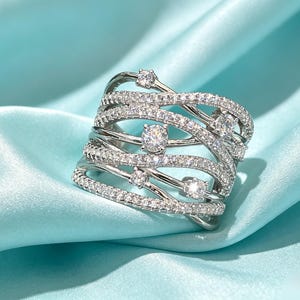 Könnte beinhalten: Ein breiter silberfarbener Ring mit mehreren überlappenden Bändern, die mit kleinen, klaren Pavé-Steinen besetzt sind. Mehrere größere, runde, im Brillantschliff geschliffene klare Steine sind in Abständen auf den sich kreuzenden Metallbändern gefasst.