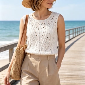 May include: A sleeveless white knit top with an intricate openwork pattern, paired with beige tailored shorts featuring a cuffed hem. A straw hat and a woven straw tote bag with brown leather straps complete the ensemble.