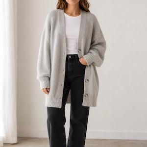 Può includere: Un cardigan in maglia grigio chiaro a maniche lunghe con scollo a V, polsini a coste e orlo a coste. Il cardigan è chiuso da quattro bottoni marroni sul davanti ed è indossato sopra una t-shirt bianca semplice con jeans neri.