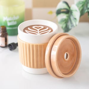 May include: A ceramic travel mug with a white interior and a ribbed, tan-coloured exterior sleeve. The mug contains a brown beverage with a white heart-shaped latte art design. A matching tan ceramic lid with a central circular hole sits beside it.