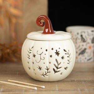 May include: A cream-coloured ceramic pumpkin-shaped candle holder with a glossy brown stem. The body features intricate cut-out patterns of leaves, stars, and swirling lines, allowing light to pass through the hollow interior of the vessel.