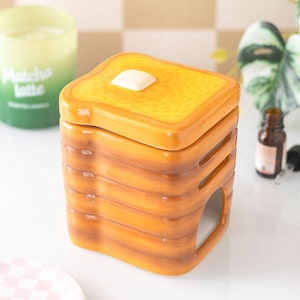 May include: A ceramic oil burner shaped like a stack of toasted bread slices. The top piece features a white square butter pat. The body has horizontal ridges resembling bread crusts and a large arched opening at the base for a tea light candle.