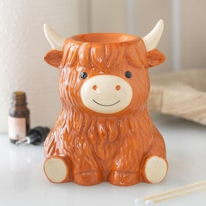 May include: A ceramic oil burner shaped like a Highland cow with a glossy orange-brown finish, cream-coloured horns, and a cream-coloured muzzle. The cow has a smiling face, black eyes, and a bowl at the top for essential oils and wax melts.