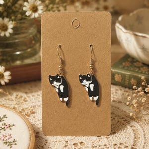 May include: A pair of dangle earrings featuring a black and white cat design. Each earring has a gold-toned metal hook and a flat, enamel-style charm depicting a cat in a hanging position. The earrings are displayed on a rectangular brown card.