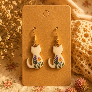 May include: A pair of drop earrings featuring white cat-shaped charms with gold-toned metal hooks. Each charm displays a colourful floral pattern on the back, including purple, yellow, and blue flowers with green stems, set against a tan card.
