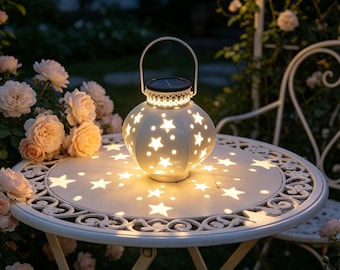 Può includere: Una lanterna sferica in metallo bianco con un motivo a stella ritagliato e una maniglia superiore. La lanterna emette una luce calda, proiettando motivi luminosi a forma di stella sulla superficie di un tavolo rotondo in metallo bianco circondato da rose.