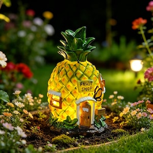 Op de afbeelding: Een ananasvormig tuinornament met een gloeiend interieur, voorzien van een bruine deur, een klein raam en een bordje met de tekst FRUIT SHOP. De buitenkant is geel en groen met gebeeldhouwde vlinderaccenten aan de zijkant van de structuur.