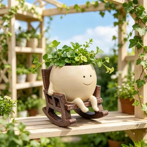 May include: A cream-colored, round ceramic planter with a simple smiling face, featuring two small arms and legs, resting in a dark brown, wood-textured miniature rocking chair, containing a lush green plant with small, rounded leaves.