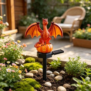 Puede incluir: Una estaca de jardín solar con un dragón naranja brillante de alas extendidas posado sobre una calabaza iluminada con cara tallada. La unidad incluye un poste de montaje negro y un pequeño panel solar rectangular en la base.