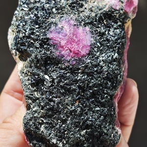 Può includere: Un esemplare minerale grezzo caratterizzato da un grande cristallo rosa magenta vibrante incastonato in una matrice scura e strutturata di orneblenda nera. La pietra presenta una superficie naturale ruvida con strutture cristalline.