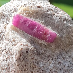Peut inclure: Un cristal magenta vibrant et allongé incrusté dans une matrice minérale brute, texturée et blanc cassé. Le cristal présente une qualité semi-translucide avec des stries naturelles visibles et une forme prismatique légèrement irrégulière.