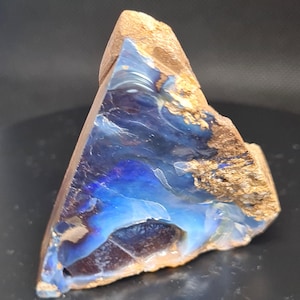 Puede incluir: Un espécimen de ópalo boulder de forma triangular que presenta una cara pulida de color azul iridiscente y violeta intenso con patrones en espiral. La piedra tiene una matriz de roca natural rugosa, texturizada y de color marrón claro.