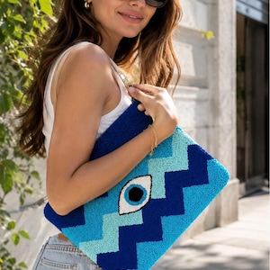 May include: A rectangular clutch bag featuring a textured, tufted surface with a blue and light blue chevron pattern. The centre of the bag displays a prominent eye motif with a white sclera, a light blue iris, and a black pupil, all in a punch needle style.