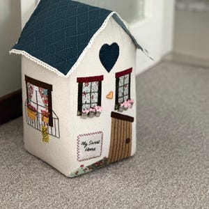 Könnte beinhalten: Ein Stoff-Türstopper in Hausform mit einem gesteppten, dunkel-türkisfarbenen Dach, weißen Wänden und einer braunen Tür. Die Vorderseite zeigt zwei Fenster mit rosa Blumen, ein dunkelblaues Herz und ein Schild mit dem Text My Sweet Home.