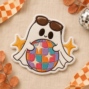 May include: An embroidered patch featuring a white ghost character wearing brown sunglasses. The ghost holds a multicoloured disco ball with a checkered pattern in shades of pink, orange, blue, and purple, accented by yellow star shapes on each side.