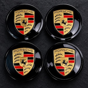 May include: Four circular black wheel center caps, each featuring the gold, red, and black Porsche crest emblem in the center. The crest includes the word PORSCHE at the top and STUTTGART in the middle, with a black horse graphic on a gold shield.