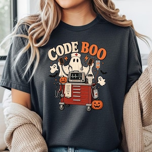 Può includere: Una t-shirt grigio scuro con una stampa grafica che riporta il testo CODE BOO. Il design mostra un fantasma vestito da infermiere, un carrello medico rosso, un monitor cardiaco, zucche e pipistrelli a tema Halloween.