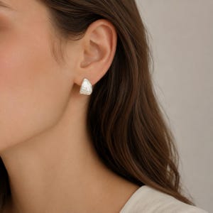 Peut inclure: Une petite boucle d'oreille à clou de couleur argentée avec une forme incurvée en demi-cercle. La surface de la boucle d'oreille présente une finition martelée texturée qui capte la lumière, créant un motif subtil, réfléchissant et irrégulier.