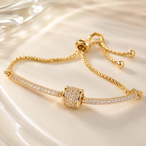 May include: A gold-toned adjustable bracelet featuring a central cylindrical bead encrusted with clear stones, flanked by two curved bars set with rows of small clear stones, connected by a delicate box chain with a sliding clasp mechanism.