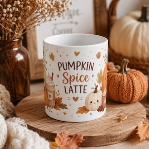 Może przedstawiać: Biały ceramiczny kubek z napisem PUMPKIN SPICE LATTE w brązowych i pomarańczowych literach. Wzór zawiera ilustracje uśmiechniętych dyń, jesiennych liści, gwiazdek, serduszek oraz napoju z bitą śmietaną w szklance.