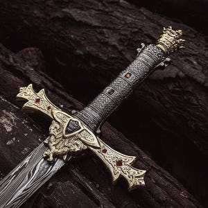 Pode incluir: Uma espada com lâmina de aço Damasco e guarda dourada com gravuras intrincadas e pequenos detalhes vermelhos. O cabo é texturizado com um padrão escuro e finalizado com um pomo dourado em formato de coroa.