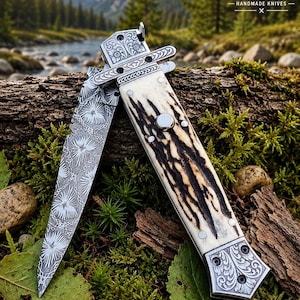 Puede incluir: Una navaja plegable con hoja de metal estampada y mango texturizado de color crema con vetas marrón oscuro. El mango tiene virolas de metal plateado con grabados florales y un mecanismo de botón circular en el centro.