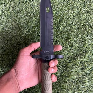 Può includere: Un coltello a lama fissa nero con dorso seghettato e un foro nella lama. L'impugnatura è verde oliva testurizzata con un pomolo nero. La lama riporta la scritta BGOK. Il coltello presenta una guardia nera che separa la lama dall'impugnatura.